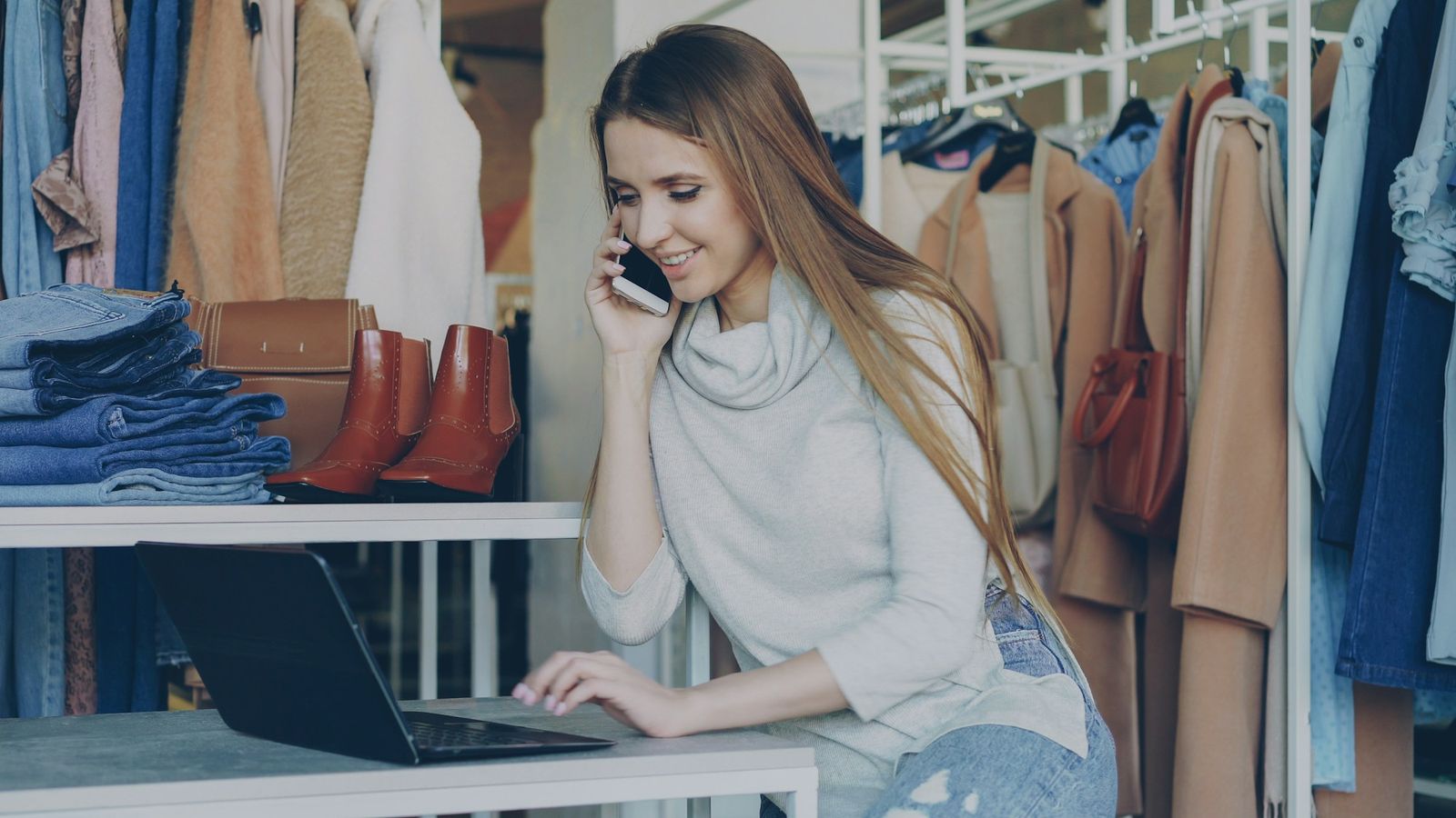 Shop owner working on her laptop and taking a customer call in her clothing store